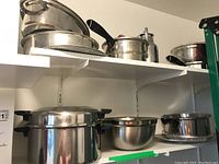 Overview of two white shelves containing multiple stainless steel pots, pans, a mixing bowl, and a metal carafe in a storage area