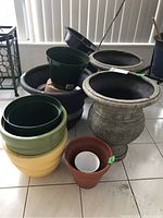 Front view of various pots and planters including green plastic nursery pots, large grey urn-shaped resin planters, yellow and light green glazed pots, and terracotta pot with plastic insert.