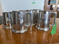 Multiple pewter steins displayed on a wooden surface, showing metallic shine and engraved designs.