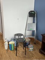 Full lot overview showing aluminum folding ladder, two black IKEA stools, two watering cans, decorative wastebasket and two tins grouped on floor against wall