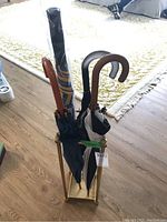 Front view of the brass umbrella stand holding four umbrellas with wooden curved handles, one umbrella covered in plastic with blue and yellow pattern visible.