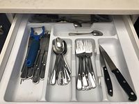 Drawer with metal flatware including forks, spoons and steak knives with black handles and a blue vegetable peeler