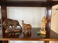 Photo of three figurines on a wooden shelf: wood buffalo on left, wood elephant in center, ceramic fisherman on right.