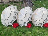 Three white crocheted lace and satin parasols and four red faux rose bouquets arranged on grass by a tree.