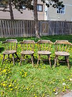 Four naturally distressed antique wooden captain's chairs arranged outdoors on grass against a wooden fence, showing front view and general condition.