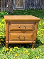 Front view of solid wood nightstand showing two drawers, brass handles, tapered legs, and medium brown finish.