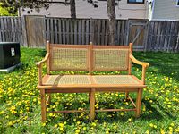 Front view of solid wood bench with woven wicker seat and backrest on a lawn with dandelions.