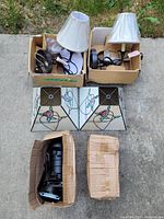 Photo showing two stained glass lampshades with rose designs, two white-lampshade wall sconces in boxes, and two industrial black spotlights in boxes, all laid out on the ground.