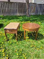 Two antique French provincial side tables placed on grass; one rectangular unfinished wood top table with carved legs and drawer, one round finished wood table with drawer.