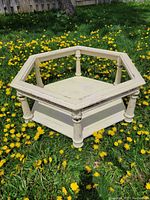 Shabby chic octagon coffee table in cream distressed paint on grass outdoors, showing spindle legs and lower shelf, missing glass top.