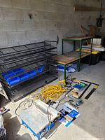 Photo shows a black wire wall shelf with multiple blue plastic parts containers, a yellow extension cord, tile saw with power cord, steel rulers, safety goggles, painter's tape, hammer, and other assorted hand tools on the floor beside the shelf and a wood and steel framed L angle workbench in the background.