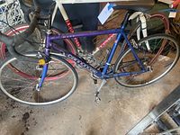 Full side view of the purple and navy Cannondale R500 road bicycle leaning against a wall in a garage. Visible pedals with clips, drop handlebars, and wheels with thin road tires.