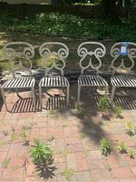 Photo showing the four vintage wrought iron scrolled dining chairs lined up outdoors on brick surface, highlighting their weathered condition and distinctive back design.