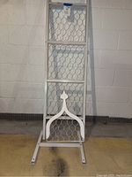 White metal garden trellis upright against a cement wall showing full height and base stand.