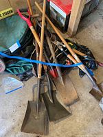Photo of gardening tools grouped on a garage floor, showing shovels with wooden handles and metal blades, pitchfork, pruners in a pile secured by a blue strap, with parts of folded lawn bags visible nearby.