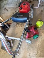 Full view showing Honda red gas lawnmower with beige grass catcher bag and metal handles. Yellow gas can and motor oil containers nearby.