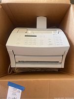 Boxed HP LaserJet 3100 laser printer seen from the top showing front keypad and output tray inside a cardboard moving box.