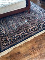 Full view of the Karastan wool rug showing deep blue border with floral designs and beige fringes. Positioned under a bed, hardwood flooring visible.
