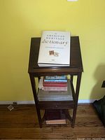 Solid wood library stand against a yellow wall, showing the top and front view with books on top and shelves below