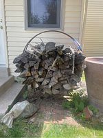 Frontal image shows the circular wrought iron wood rack loaded with firewood (wood not included in sale) on a brick patio next to house siding. Partial bag of gravel visible on ground adjacent to rack.