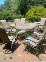 Full view of rectangular outdoor dining table with six stationary chairs and two swivel armchairs arranged around it on a brick patio with overgrown weeds.