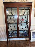 Front view of vintage dark wood china cabinet with glass front doors featuring geometric wood design and fabric-lined shelves inside with displayed glassware.
