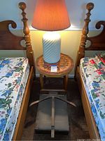Photo showing the white swirl glass lamp with beige pleated shade on top of the octagonal side table, between two wooden beds with floral bedding. Lamp is shown turned on.