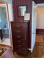 Full view of tall lingerie chest against patterned wallpaper next to doors, showing overall height and front view with ornate brass pulls.