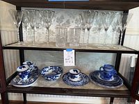 Shelf showing 11 long-stemmed wine glasses and 4 highball drinking glasses on the top shelf, with blue and white Waldorf of England dishes on the shelves below