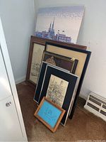 Seven framed wall art pieces stacked upright against wall. Includes canvas cityscape and various framed architectural sketches.