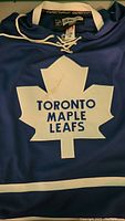 Close-up photo of front blue Toronto Maple Leafs jersey showing stained white maple leaf logo with text 'TORONTO MAPLE LEAFS'. Laced collar visible.