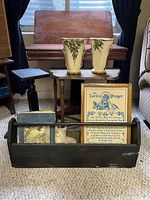 Photo showing the antique wood flip top table, two small wooden stands, McCoy pottery vases, a black wooden toolbox with a vintage book and cat figurine, and a framed needlepoint of The Lord's Prayer.