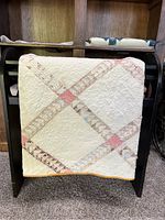 Antique hand pieced quilt draped over a dark wooden rack showing the lattice pattern of muted pastel fabric strips on cream background and gold binding at the edge.