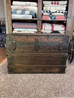 Front view of an antique wooden trunk with metal corner reinforcements, black metal bands, and two front latches. The trunk is worn and weathered.