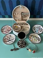 Photo showing multiple trays of jewel pops with various colorful stones, open jewelry storage case with compartments holding additional jewel pops and earring components, silver bangle bracelet, and a small black circular case.