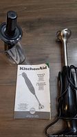 KitchenAid immersion blender main unit, blending container with lid, and product manual on wood surface