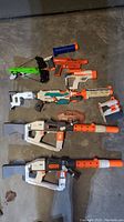 Overview of eight assorted Nerf dart blasters and accessories laid on concrete floor.