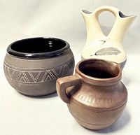 Three signed pottery pieces together: a black and grey bowl with geometric patterns, a cream double-spouted vase with feather designs, and a small brown pot with handle.
