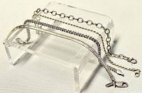 Four silver bracelets displayed on a clear stand showing different chain styles and clasps.