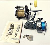 Photo of two fishing reels: Compac 100 Interceptor II spinning reel with blue line, Penn Peer #309 conventional reel with yellow handle and yellow line, alongside a small metal part and a Penn P-38 manual.