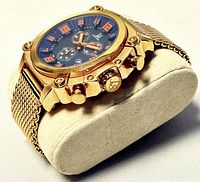 Close-up angled view of the gold-tone Joshua & Sons Swiss chronograph watch on a soft pillow with visible orange hands and numerals.