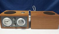 Front view of the TIVOLI Audio Model 3 pair, showing the analog clock radio and control panel on the primary speaker and the adjacent secondary speaker with wood casing and speaker grill.