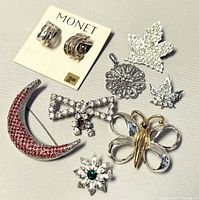 Monet clip-on earrings on card and six different brooches including a red rhinestone crescent moon, rhinestone bow, maple leaf, floral pendant, small leaf, and butterfly.