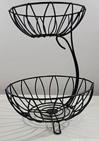 Two angled photos of a black 2 tier countertop fruit basket made of iron wire with leaf shaped design elements. Shows overall structure and sizing.