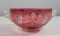 Side view showing cranberry crystal bowl with clear glass handles, detailed enamel painted floral motifs, and gilded scrolling patterns.
