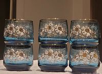 Front view of 6 stacked teal blue Dominion glass tumblers with white and gold floral decoration, showing detailed design and color.