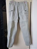 Front view of a pair of light gray men's utility pants showcasing zip-off legs and multiple front and side pockets made from ripstop fabric.