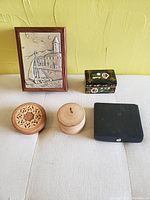 Photo of the four trinket boxes - three wood and one leather covered. Shown closed with various lid styles and a framed relief art piece in background.
