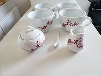 Photo of 4 large white mugs each with red leaf pattern, along with a sugar bowl with lid, creamer, and spoon on a white surface