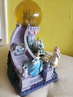 Angled view of Disney Cinderella musical staircase figurine showing Cinderella at bottom of lavender stairs, pumpkin carriage, white horses with pink feather, yellow globe with castle inside.